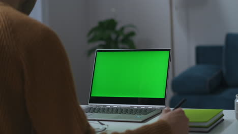 Laptop-Mit-Grünem-Bildschirm-Auf-Dem-Arbeitstisch-Im-Home-Office,-Blick-Durch-Die-Schulter-Eines-Mannes,-Der-Remote-Arbeitet-Und-Freiberuflich-Videokonferenzen-Abhält